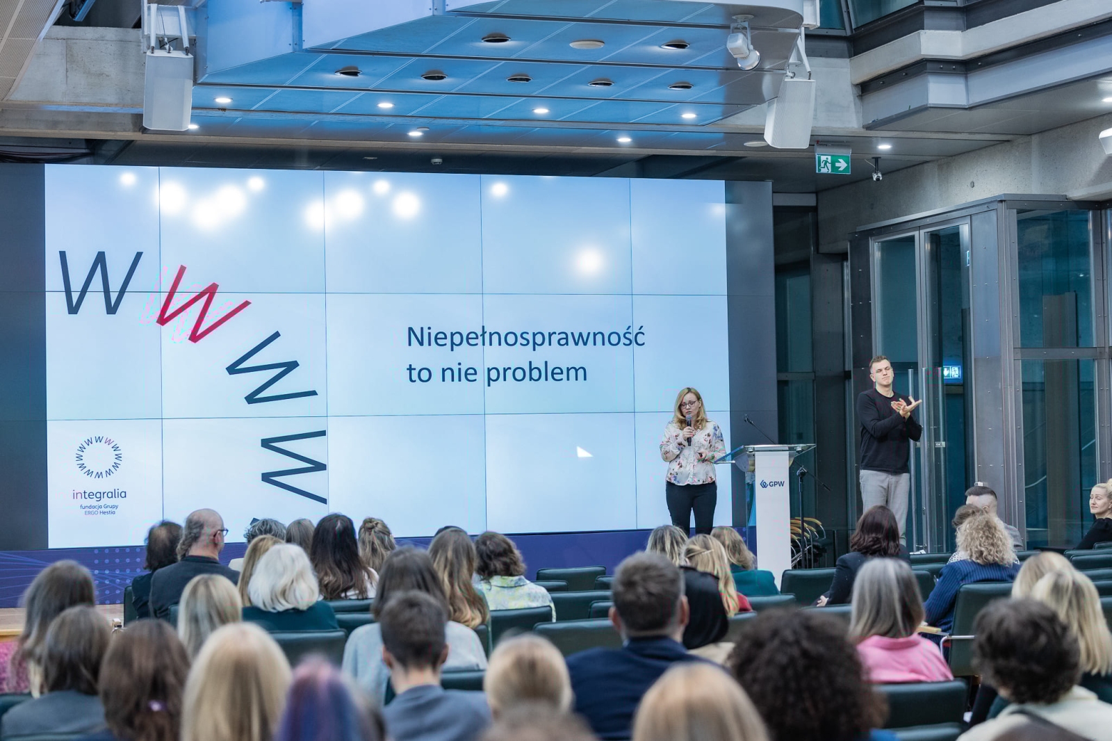 A speaker and sign language interpreter are giving a presentation in a conference room. Behind them, on a screen, is the slogan: “Disability is not a problem.” The audience is seated facing the stage.