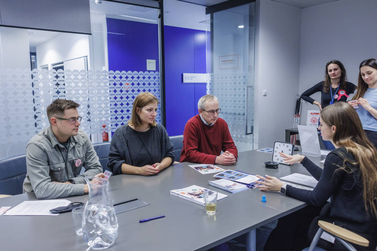 Spotkanie warsztatowe przy stole konferencyjnym. Uczestnicy rozmawiają z prowadzącą o doświadczeniach z ćwiczeń symulacyjnych i o barierach, które mogą pojawiać się w środowisku pracy.