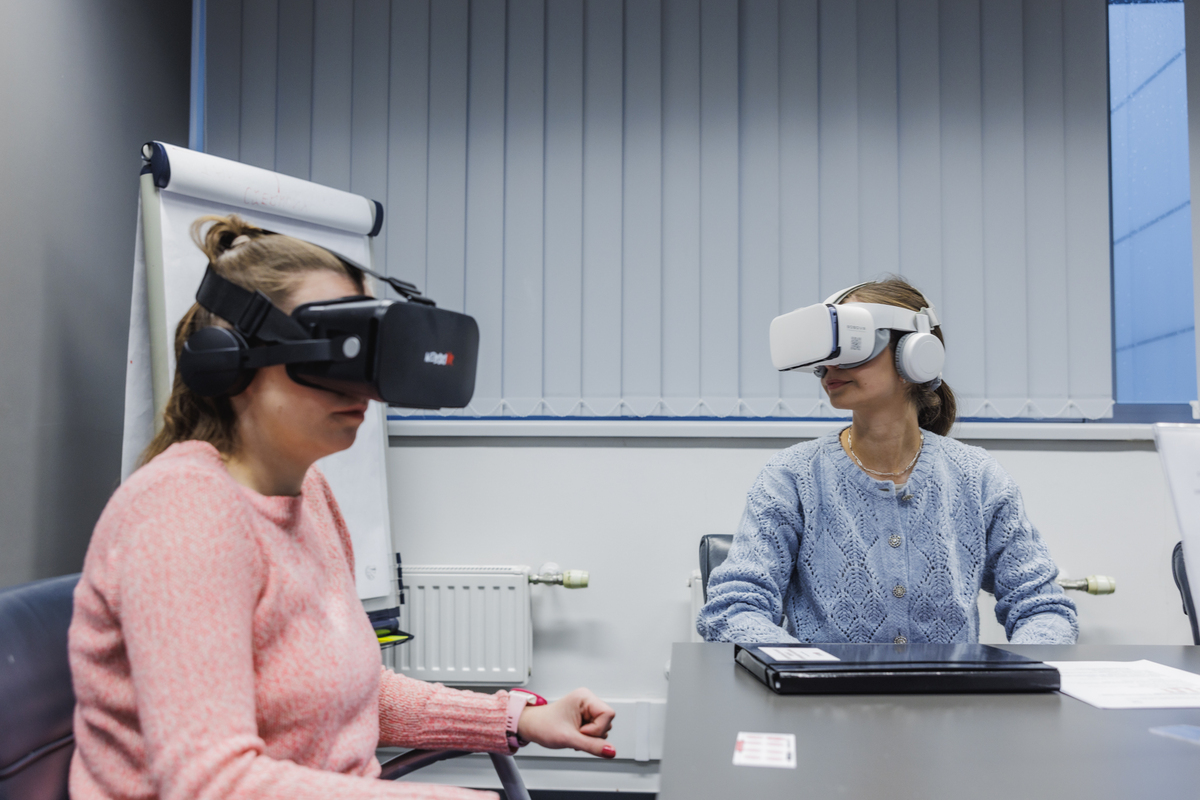 Two women sit at a table wearing VR goggles and headphones. They are practicing a virtual simulation, with a blind and a flipchart in the background. They are focused on the task at hand.