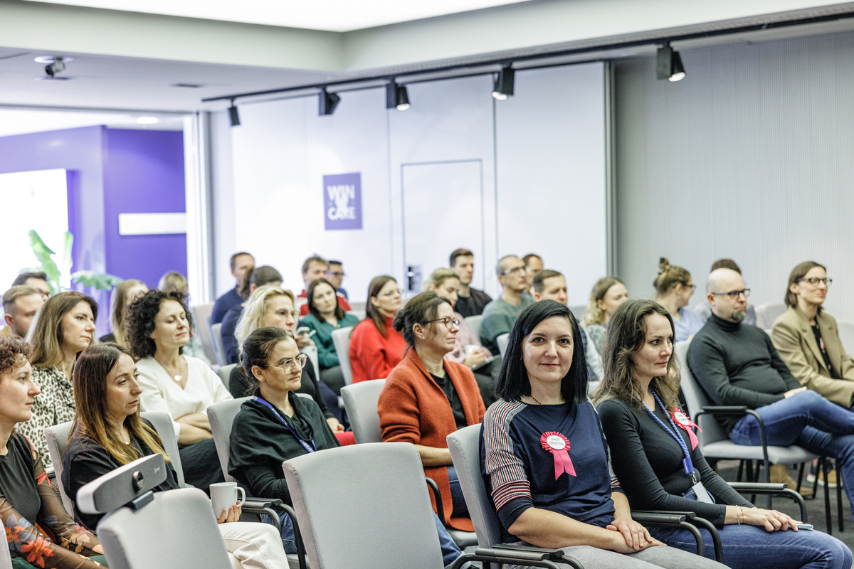 Audience at an educational event. Participants are seated in rows, listening to a presentation. Some wear rosettes reading “Change your perspective”.