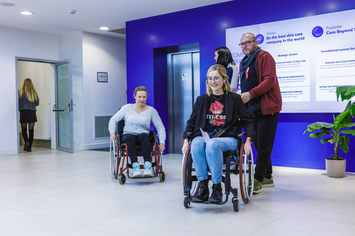 Two women in wheelchairs ride through the hallway, smiling. Accompanying them is a man with a “Change perspective” badge. In the background is a plaque with the company's mission statement.