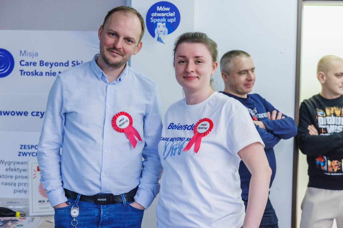A woman and a man with “Change your perspective” pins stand together and look into the lens with a smile. In the background are event participants and company posters.