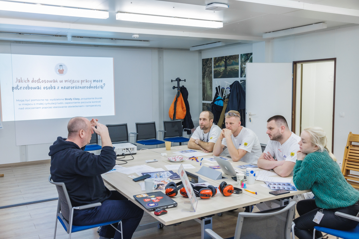 A man is having a conversation with a group of five people at a table. A slide about workplace adjustments is displayed on the screen.