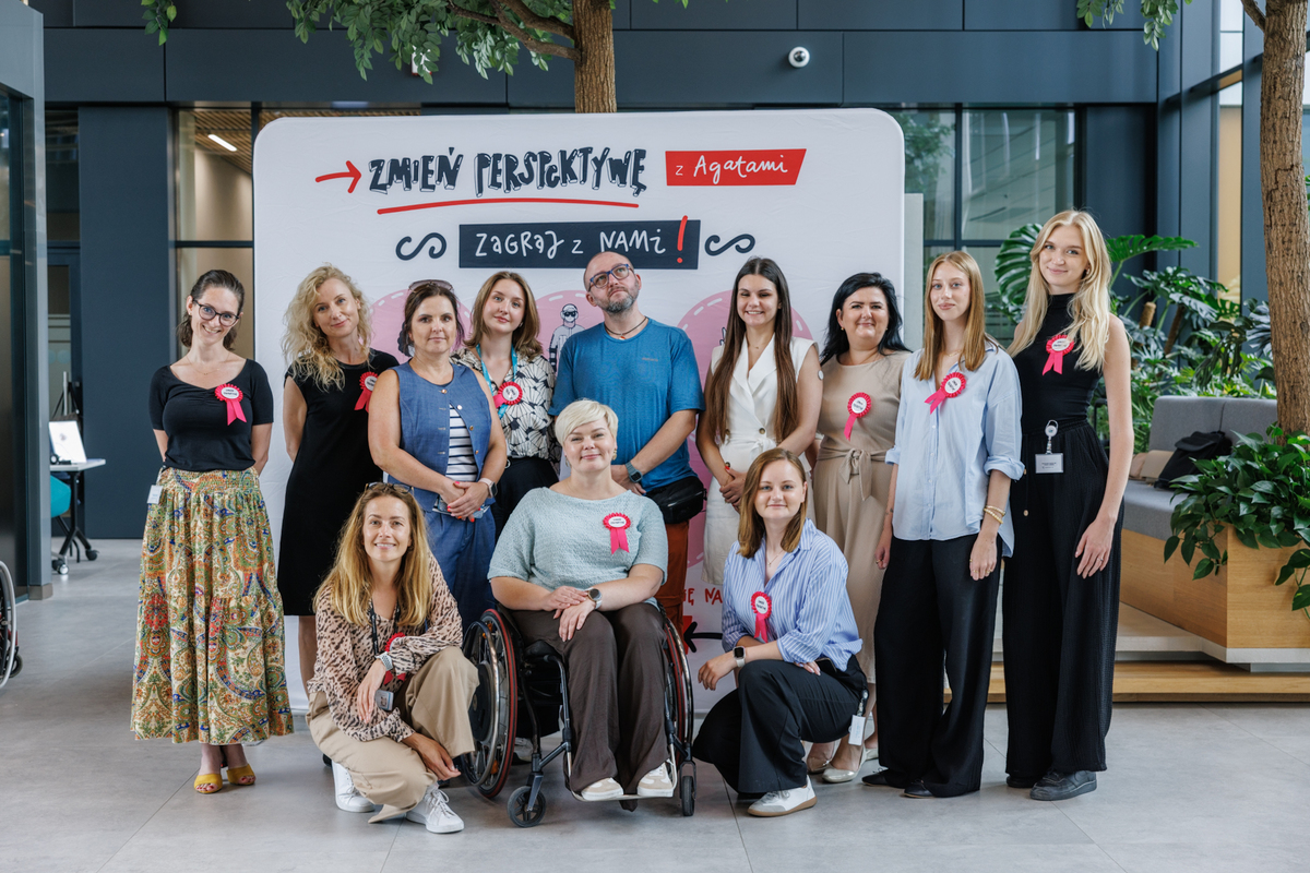 Eleven people pose together in front of the “Change your perspective with Agata” board. In the center is a woman in a wheelchair.