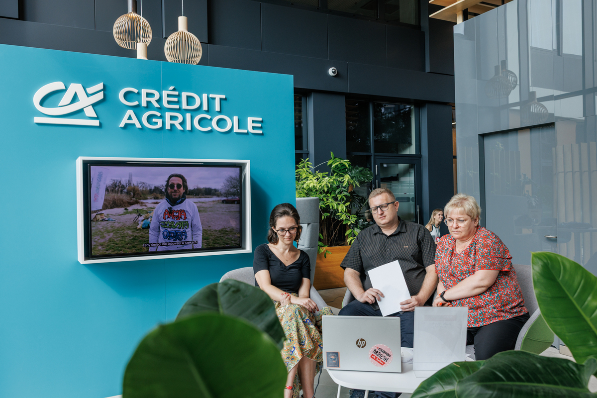 Three people are sitting at a table in the Credit Agricole space. In front of them is a laptop and documents, with a screen displaying the bank's logo and a video in the background.