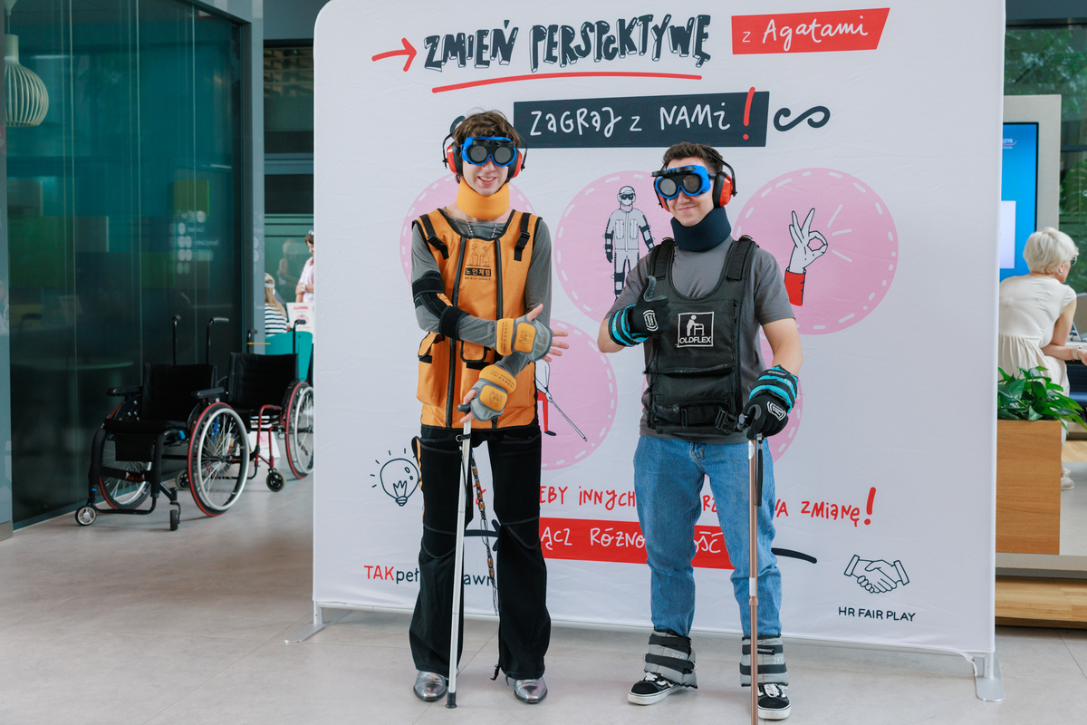 Two men in old age suits pose in front of a sign reading “Change your perspective with Agata.” Wheelchairs are visible in the background.