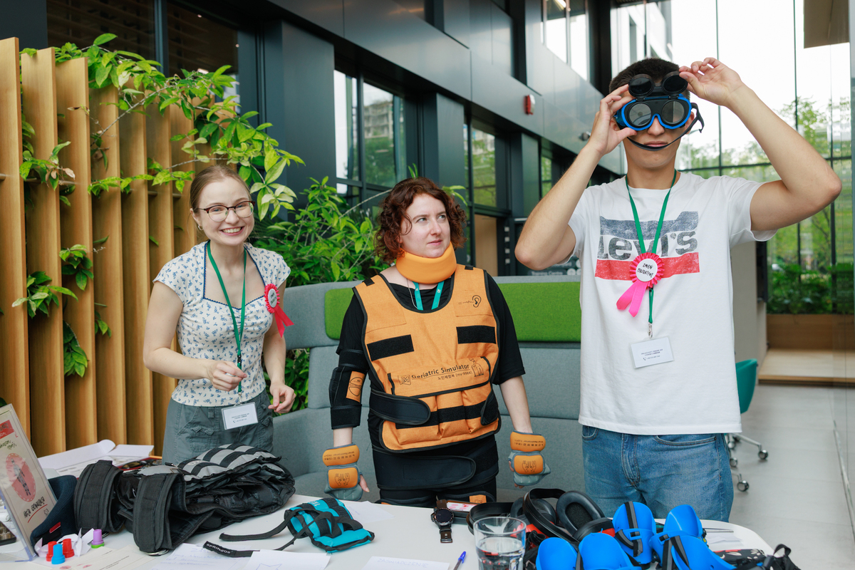 Three people at the table are testing accessibility equipment. One is wearing an aging suit, another is wearing goggles that restrict vision.