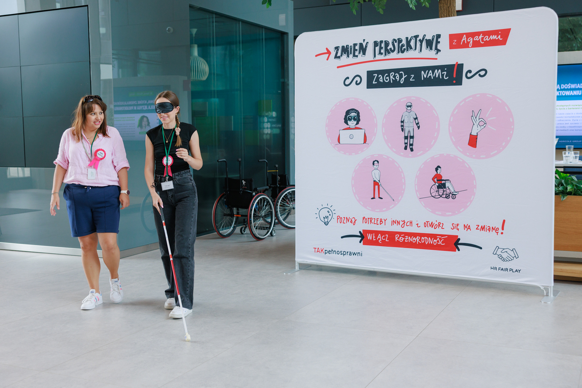 Two women are participating in an exercise with a white cane. One has a blindfold on, the other is assisting. In the background, there is a board that says “Change your perspective.”