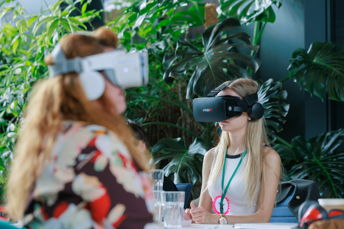 Two women participate in an exercise using VR goggles. They sit at a table surrounded by plants.