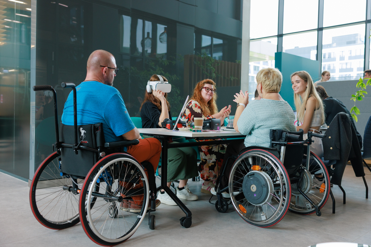 A group of people, including two in wheelchairs, are talking at a table. One of the participants is wearing VR goggles.