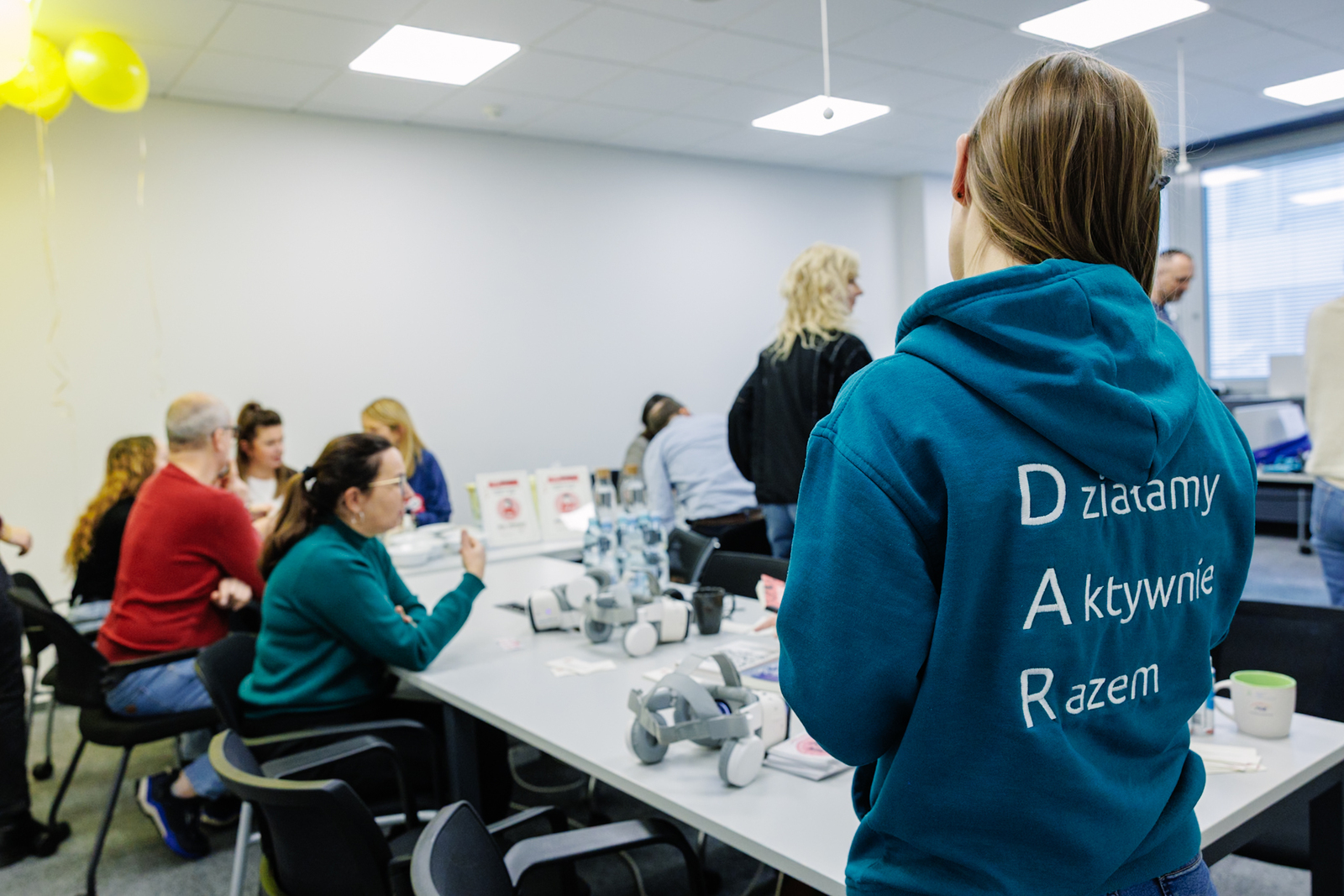W tle kilka osób rozmawia przy stole w sali szkoleniowej. Na stole google VR i materiały edukacyjne. Na pierwszym planie osoba odwrócona tyłem, w zielonej bluzie z napisem „Działamy Aktywnie Razem”. 