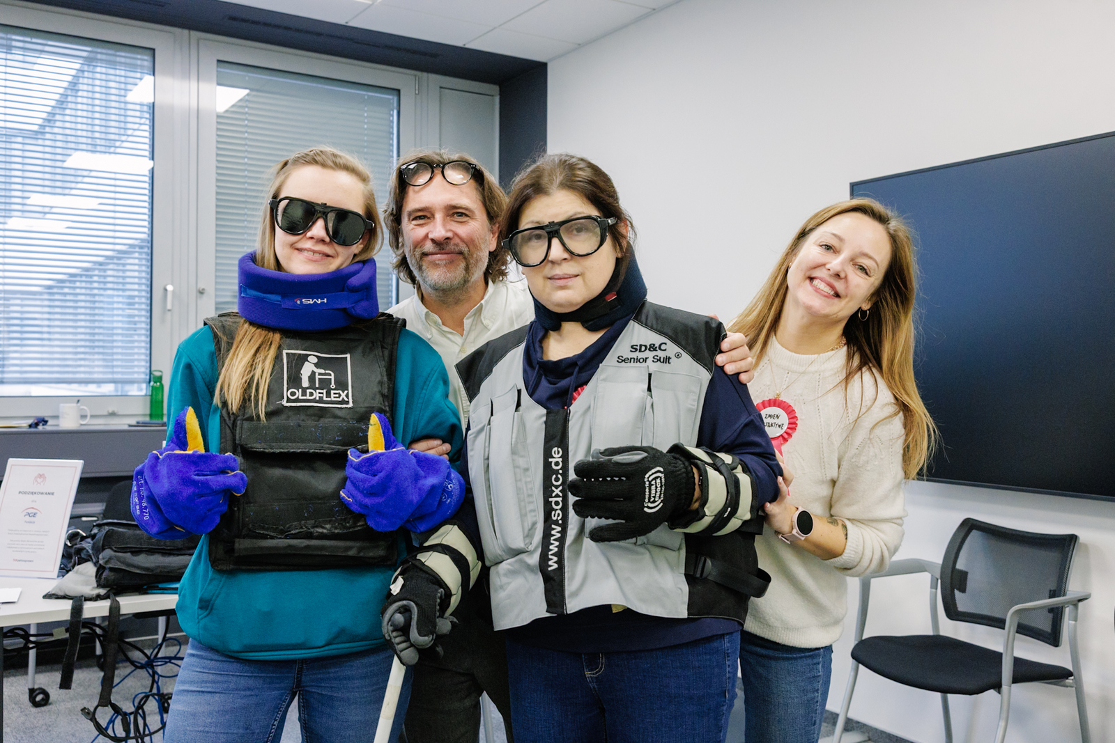 Cztery osoby, trzy kobiety i mężczyzna, stoją obok siebie w sali biurowej. Dwie mają na sobie kombinezony symulujące ograniczenia wynikające z wieku. Są uśmiechnięci. W tle ekran telewizora i krzesła biurowe.