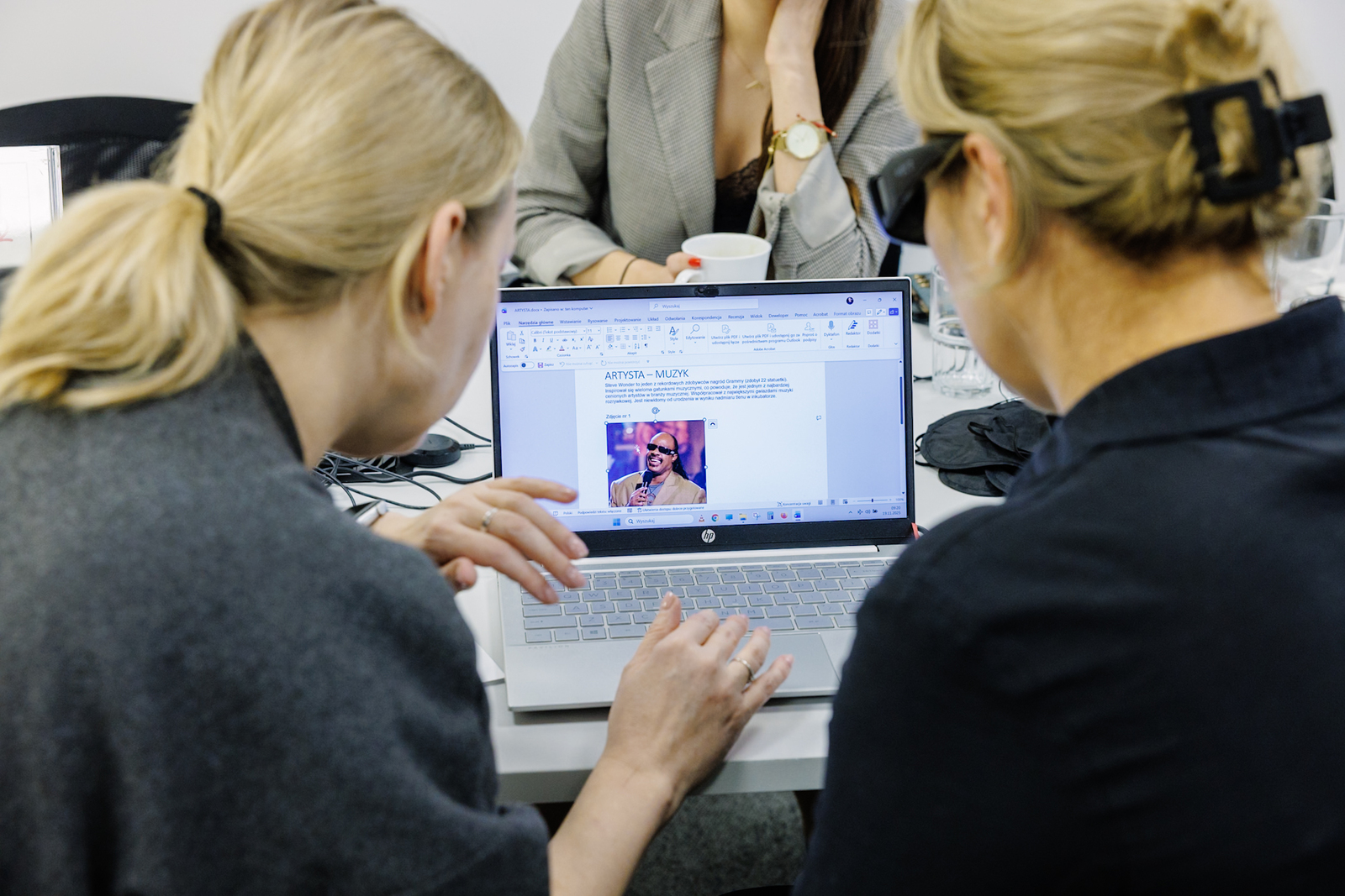 Trzy osoby siedzą przy stole, Dwie z nich wspólnie pracują przy laptopie.  Na ekranie materiał edukacyjny o niepełnosprawnościach wzroku. Trzecia osoba siedzi na wprost, trzymając w ręku kubek.