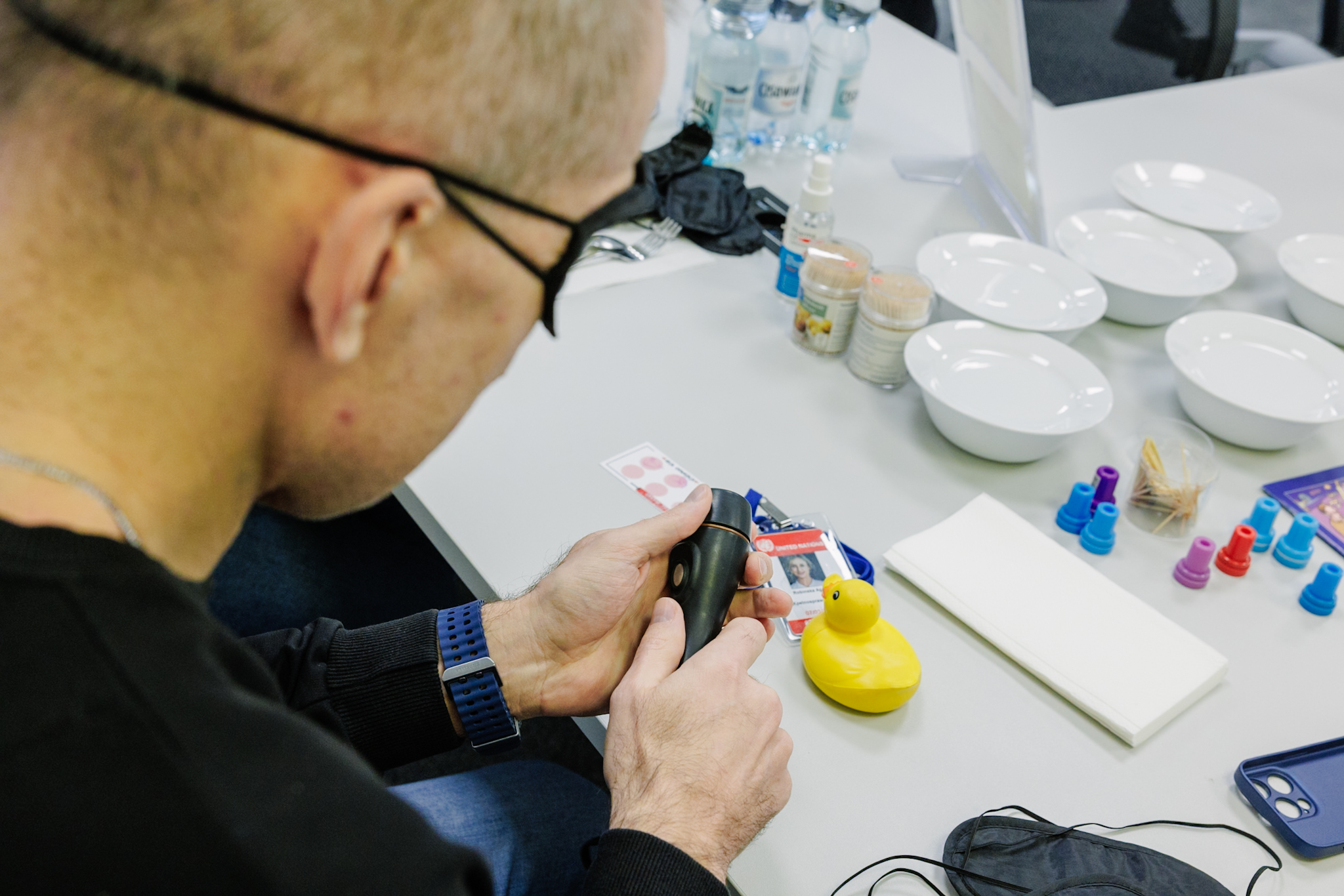 Osoba siedzi przy stole i trzyma w dłoniach niewielkie urządzenie do ćwiczeń sensorycznych w doświadczeniu braku wzroku. Na blacie miski, produkty spożywcze, karty i drobne akcesoria używane do ćwiczeń.