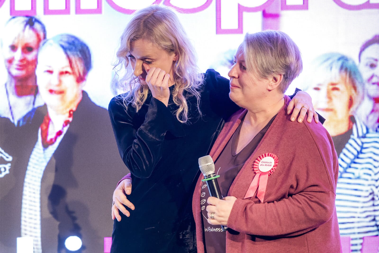 Agata Robińska and Anna Nowak stand side by side on stage. Agata wipes away a tear, Anna holds the microphone. In the background, a screen displays images of the participants in the “Breath for Moms” project.