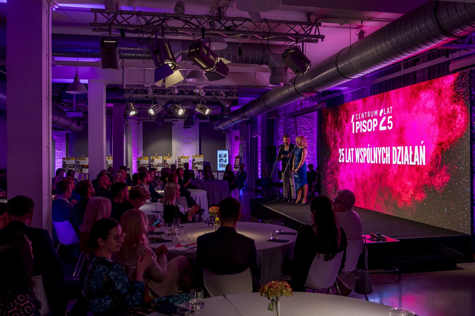 View of the hall during the Greater Poland Region Social Award gala. Participants are seated at round tables, with two hosts on stage and a screen displaying the words “25 years of joint action.”