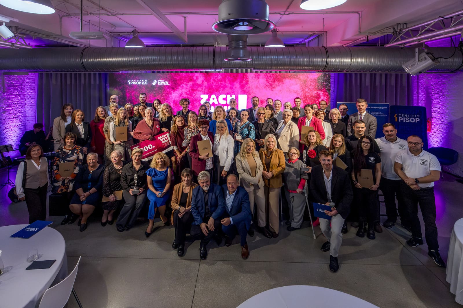 Group photo of participants at the Wielkopolska Social Award gala. Several dozen people pose in front of a screen with the word “Zachwytniki” and the logos of PISOP and the NIWA Foundation.