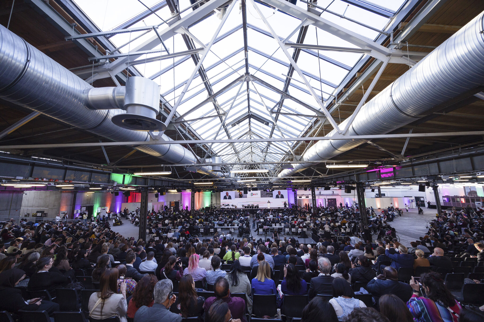 Interior of the spacious Global Disability Summit hall with a glass ceiling and colorful lighting. Hundreds of attendees face the stage with large screens and speakers.