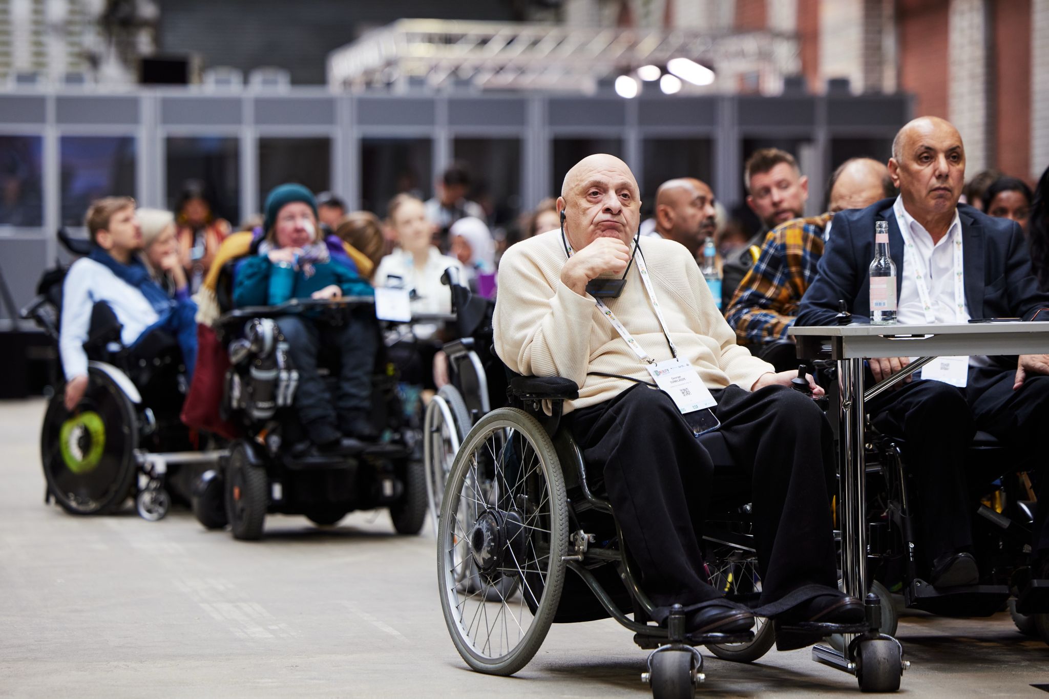 Audience during the Global Disability Summit. In the foreground, an older man and several wheelchair users are focused and listening. The background shows the hall and interpreter booths.