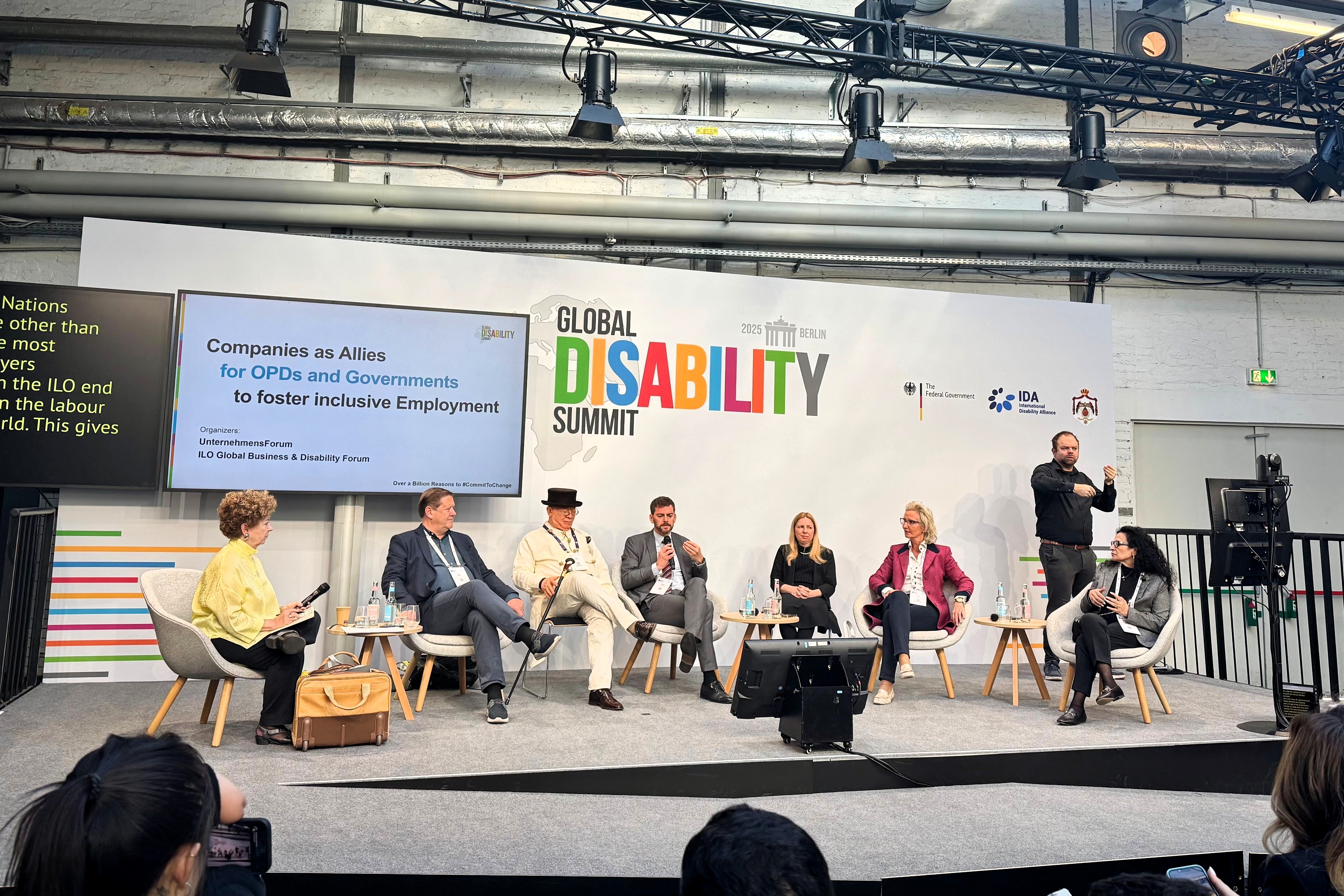 Panel discussion at the Global Disability Summit. Seven speakers sit at a table with microphones. A man in the center gestures with open arms, while others clap and smile.