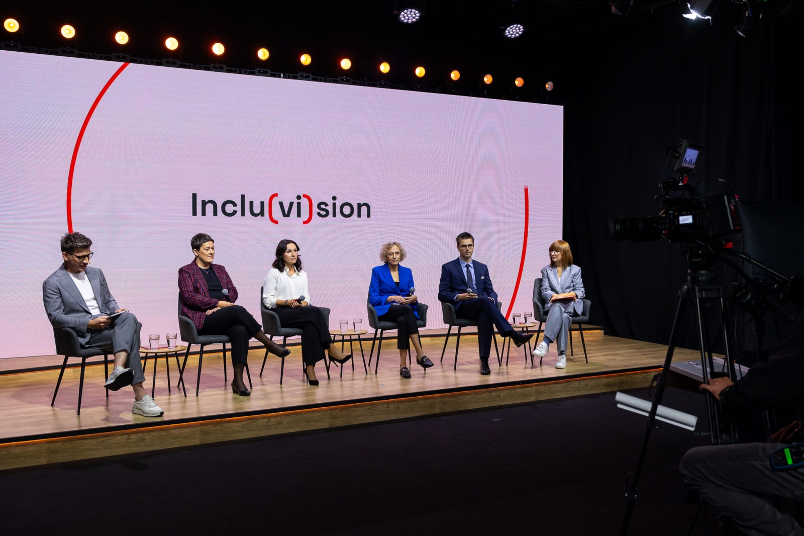 Six panelists on stage during the Inclu(vi)sion conference. They are all seated on chairs in front of a large screen displaying the event logo. A camera and stage elements are visible to the side.