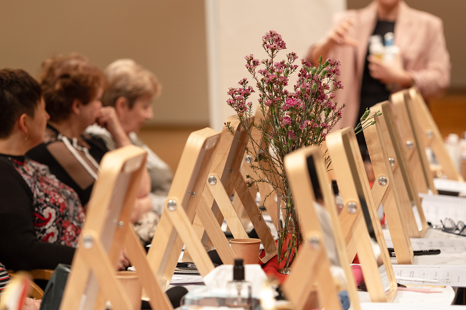 Stoły z lustrami, kosmetykami i bukietem kwiatów podczas warsztatów makijażu w Solar Company. W tle uczestniczki projektu „Oddech dla Mam” poznają techniki pielęgnacji.