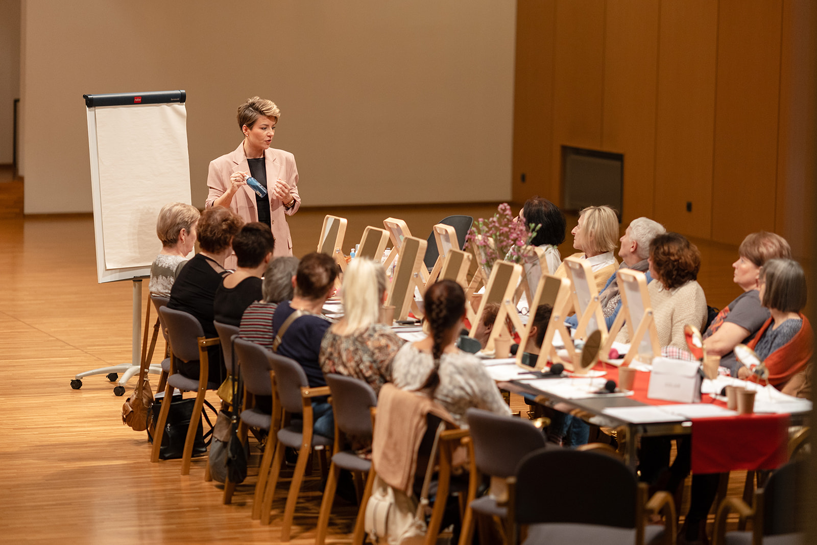 Prowadząca w różowej marynarce prezentuje kosmetyki uczestniczkom warsztatów makijażu w Solar Company. Kobiety siedzą przy stołach z lustrami w ramach projektu „Oddech dla Mam”.