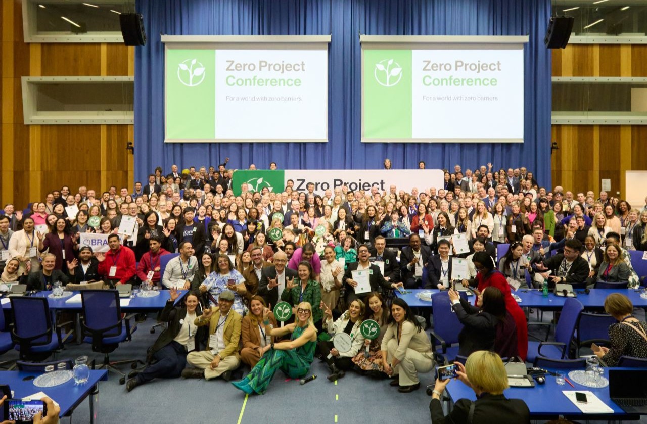 Group photo of Zero Project Conference participants in a large hall with blue curtains and event banners. Hundreds of people face the camera, many holding certificates and nameplates.